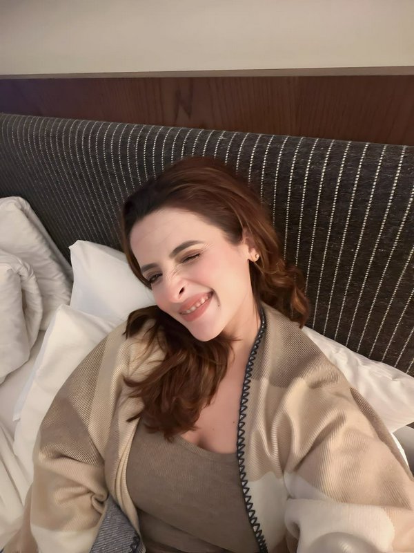 Smiling winking woman lounging in bed, wrapped in a neutral checkered shawl, propped on white pil...