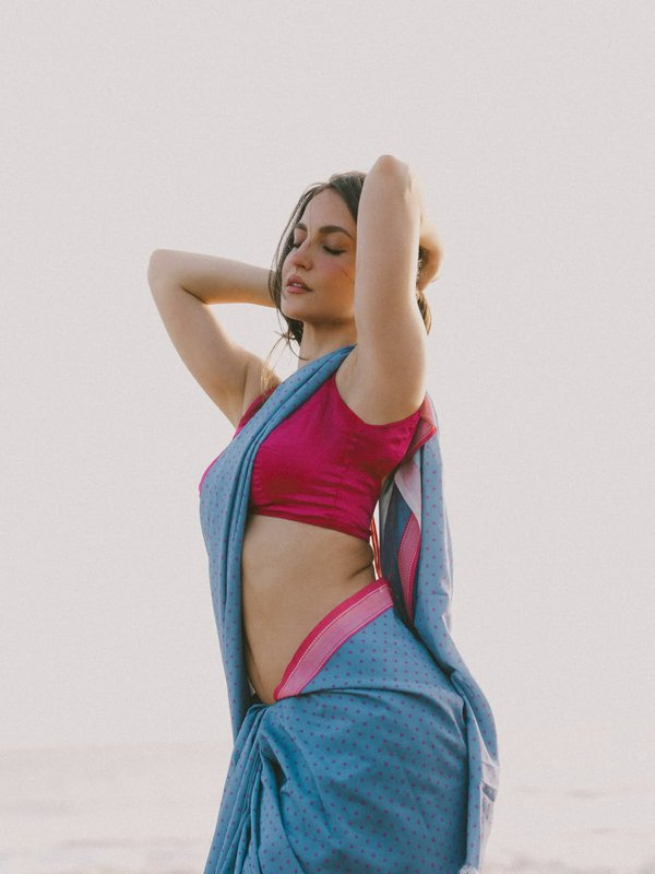 Young woman in blue polka dot saree and magenta blouse stands with arms raised, eyes closed, agai...