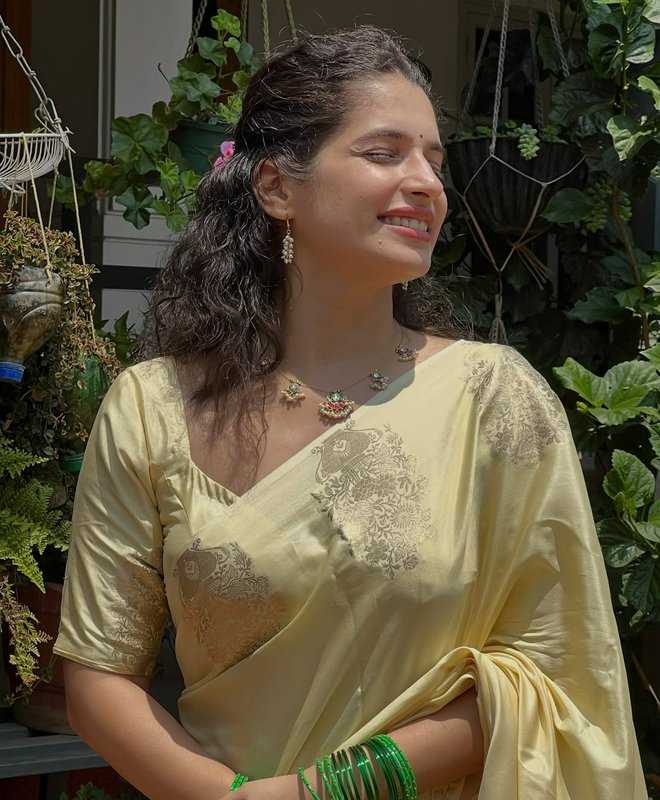 Smiling South Asian woman in pale gold silk saree stands sunlit on a plant-filled balcony, eyes c...