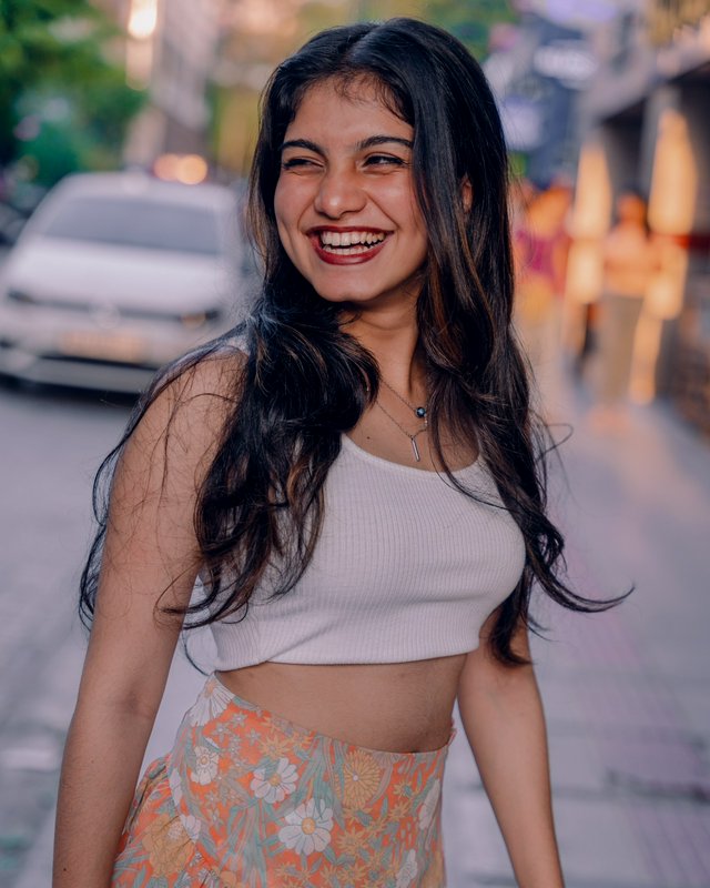 Laughing young South Asian woman with long wavy dark hair stands on a city sidewalk at dusk in ca...
