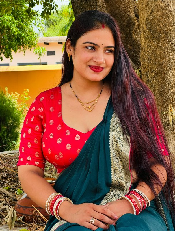 Smiling Indian woman in a teal silk saree and red blouse sits outdoors next to a tree, wearing tr...