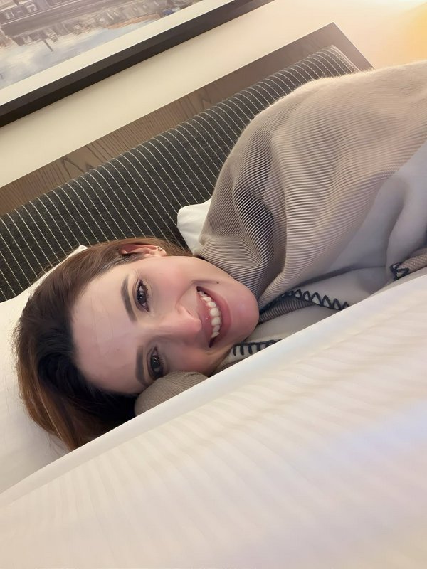 Cheerful grinning woman wrapped in a beige knit blanket lays comfortably in a bed taking a casual...