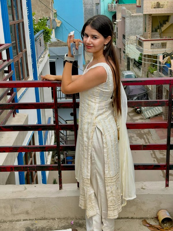 Young woman in cream embroidered Indian salwar suit poses on a residential rooftop balcony holdin...