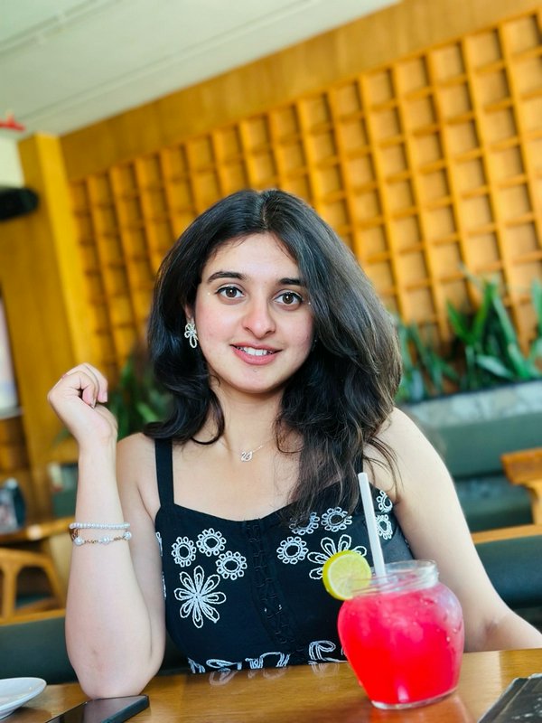 Smiling young woman seated at a cozy cafe table with a bright pink garnished iced mocktail, warm ...