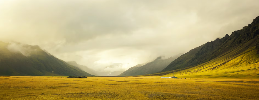 Panoramic remote mountain valley with golden grassy plains, misty jagged peaks, and tiny isolated structures under moody g...