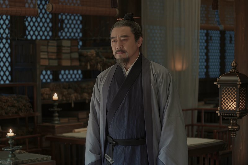 Older bearded Chinese man in traditional hanfu standing in a dim candlelit historical study