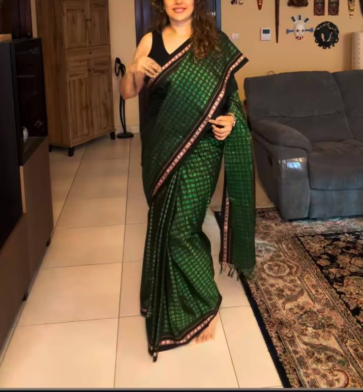 Woman modeling a dark green checkered silk saree with decorative border, standing in a cozy home ...