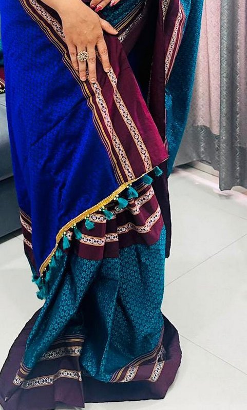 Close-up of a woman displaying a woven teal and royal blue saree with maroon border and teal tass...