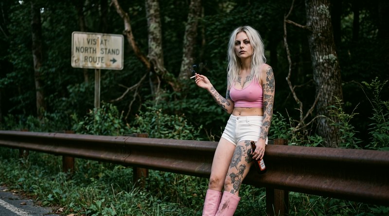 Heavily tattooed platinum blonde woman leans on a forest roadside guardrail holding a cigarette a...