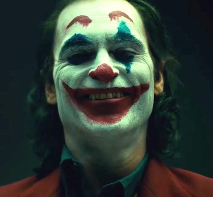Close-up portrait of Joaquin Phoenix as Joker grinning in smudged clown makeup against a dark moo...