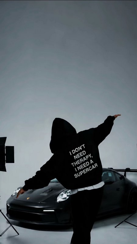 Back view of hooded person posing with outstretched arms next to a glossy black Porsche supercar ...