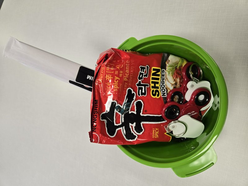 Overhead view of green instant noodle bowl holding Shin Ramyun pack, fidget spinners, and chopsti...