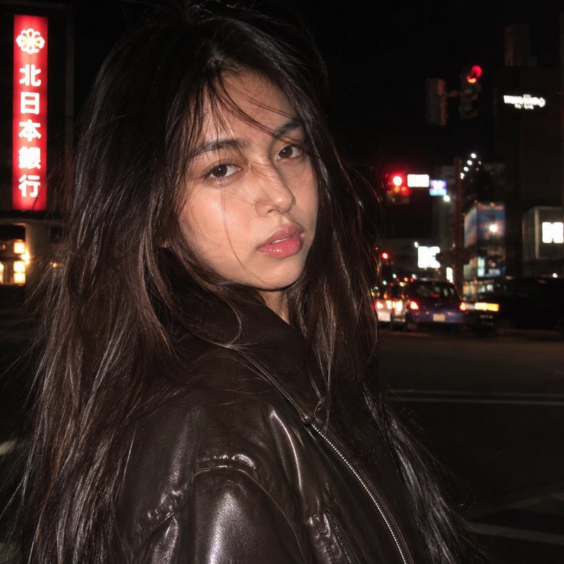 Candid portrait of a young woman in a brown leather jacket standing on a neon-lit Japanese city street at night.