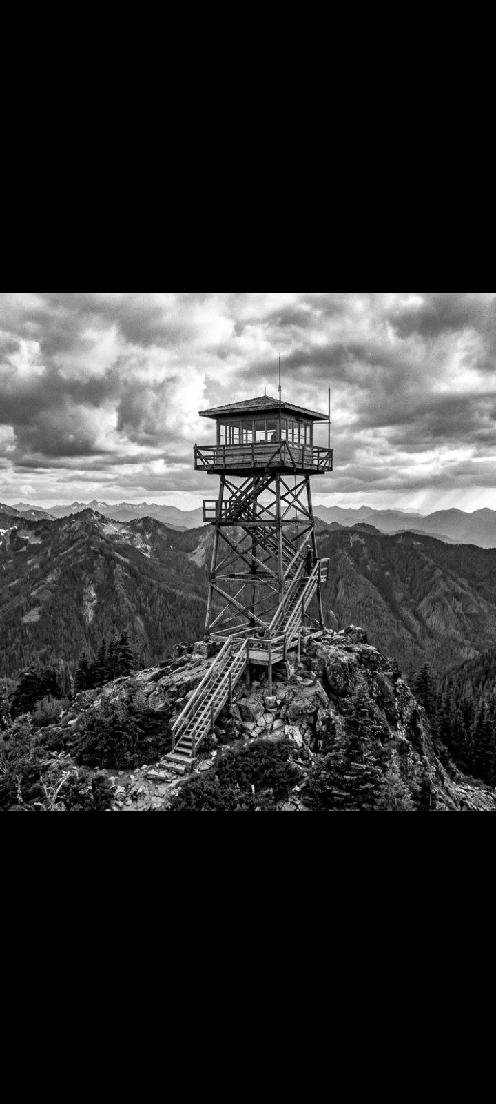 Wooden fire lookout tower perched on a rocky mountain peak under dramatic cloudy skies
