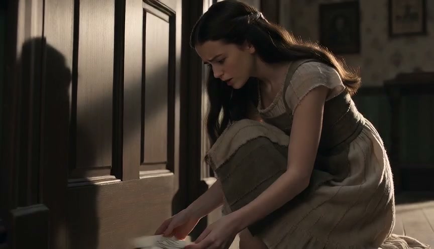 Young woman in vintage period dress kneels to read a handwritten letter beside a paneled wooden door