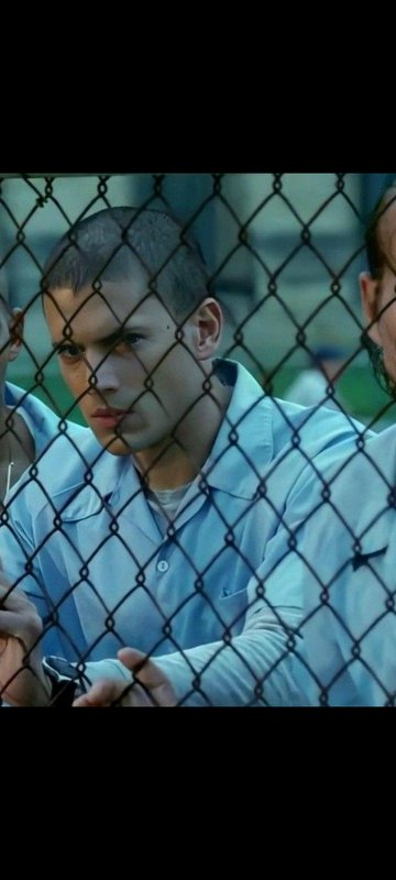 Close up of Michael Scofield from Prison Break standing behind a chain link prison fence with a f...
