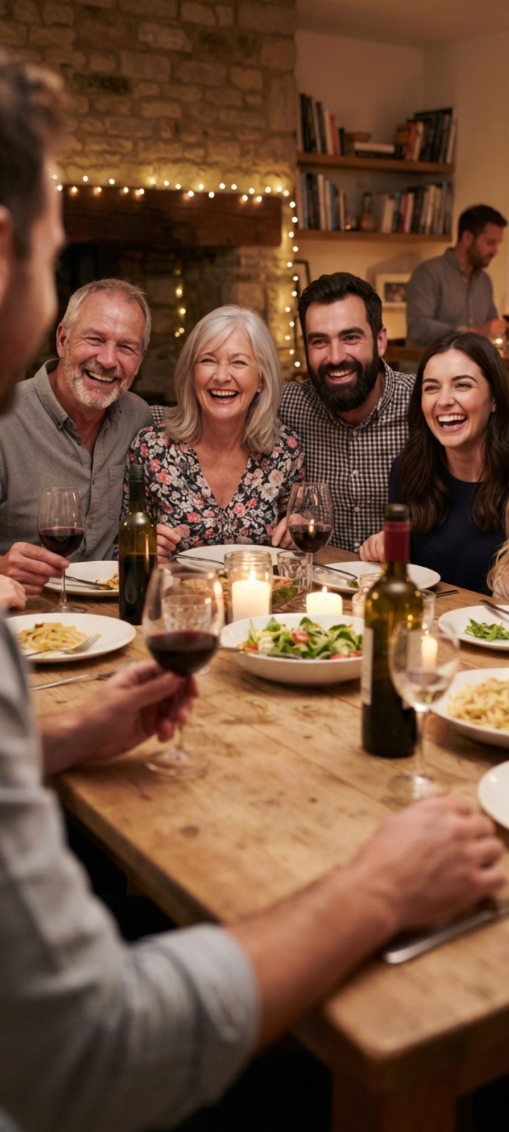 [Joyful multi-generational group laughs together around a candlelit rustic dinner table at a cozy home dinner party with w...