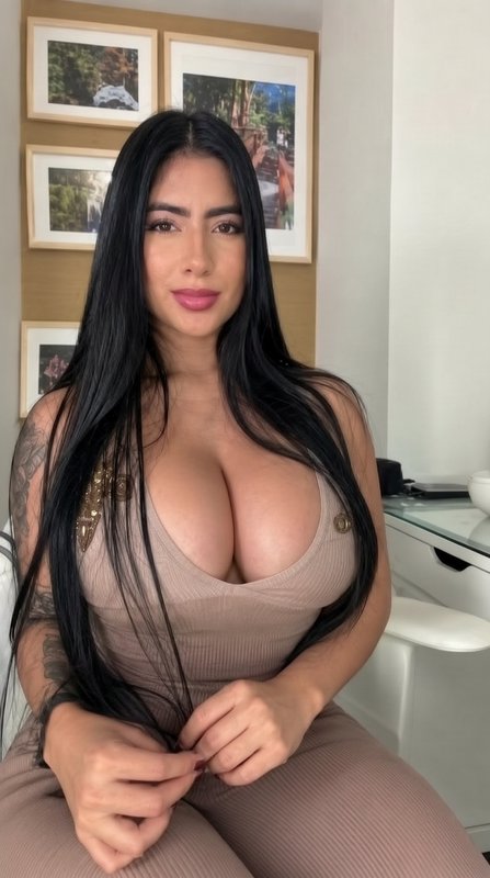 Casual indoor portrait of a woman with long straight black hair, tattooed arm, seated in a bright...