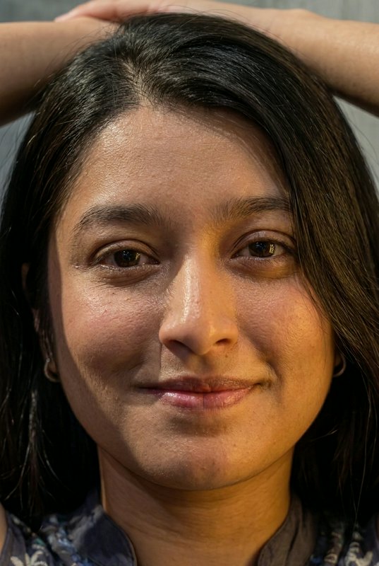 Close-up portrait of a South Asian woman with dark hair, soft smile, arms resting above her head ...