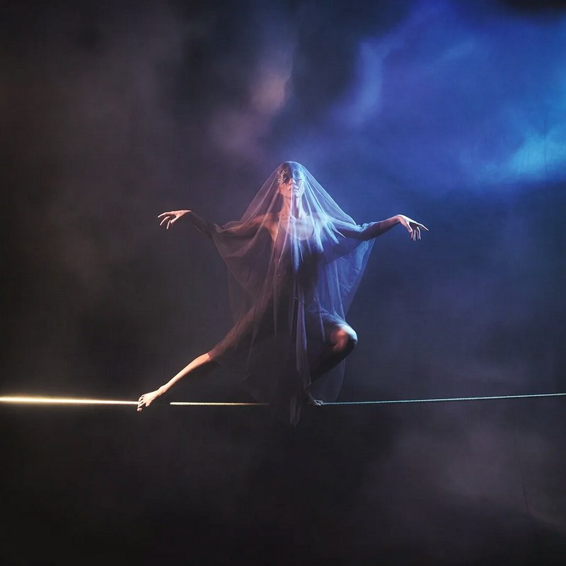 A dancer on a tightrope draped in a translucent veil under moody blue stage lighting.