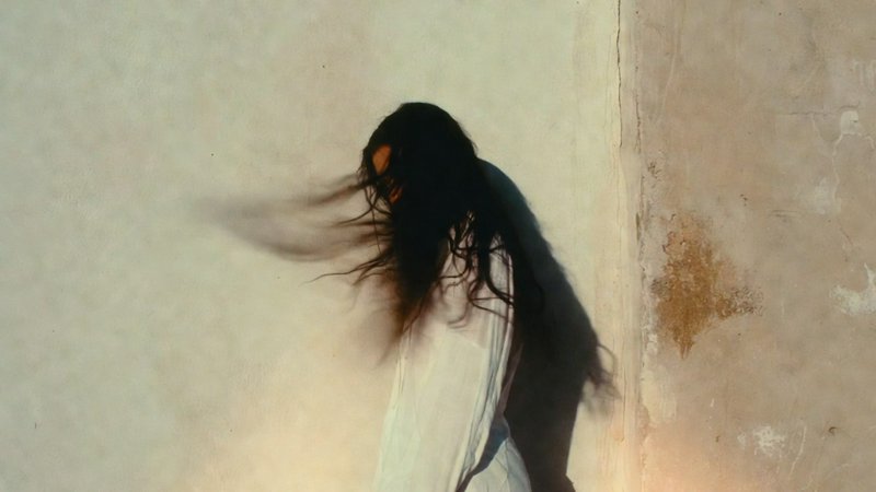 Blurred figure of a person with long hair leaning against a beige wall, motion effect.