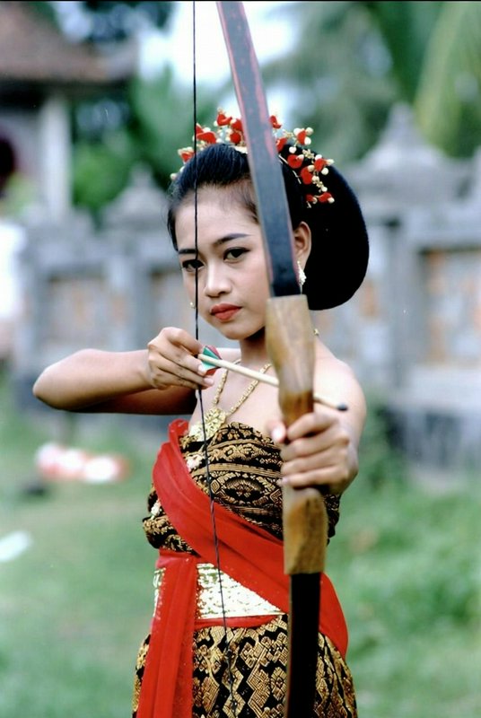 Young Balinese woman in traditional ceremonial dress aims a wooden bow on grassy temple grounds.