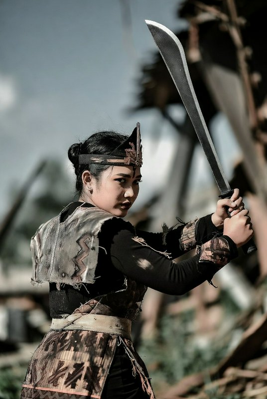 Female Southeast Asian warrior in traditional tribal armor holds a curved sword in a defensive co...