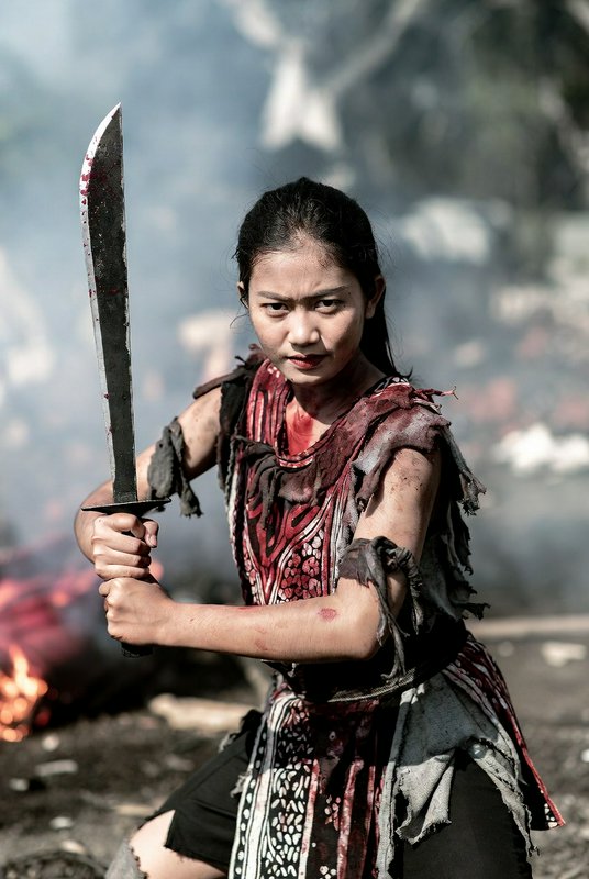 Fierce young female warrior holds a bloodied machete in combat stance on a smoky burning battlefi...