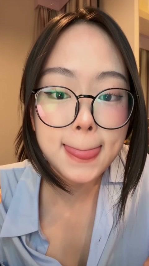 Closeup selfie of young woman with round black glasses making a playful face in a warm indoor hom...