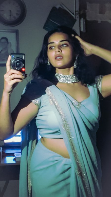 Woman in pale blue embroidered saree with silver jewelry takes mirror selfie with a compact digit...