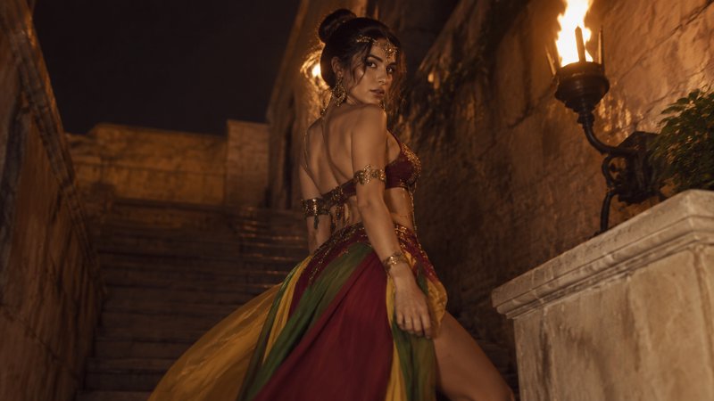 Woman in ornate fantasy costume glances back while standing on a torch-lit stone castle staircase at night.