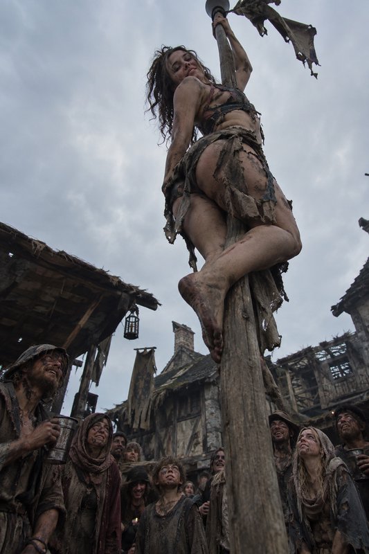 Low angle shot of a ragged, dirt-streaked woman on a tall wooden pole as medieval villagers look ...