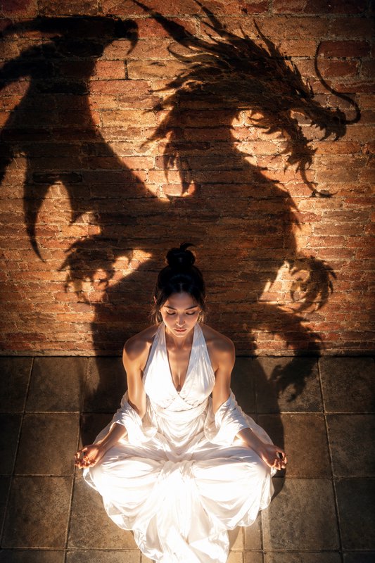 Woman meditating cross-legged with a large intricate dragon shadow cast on the brick wall behind her