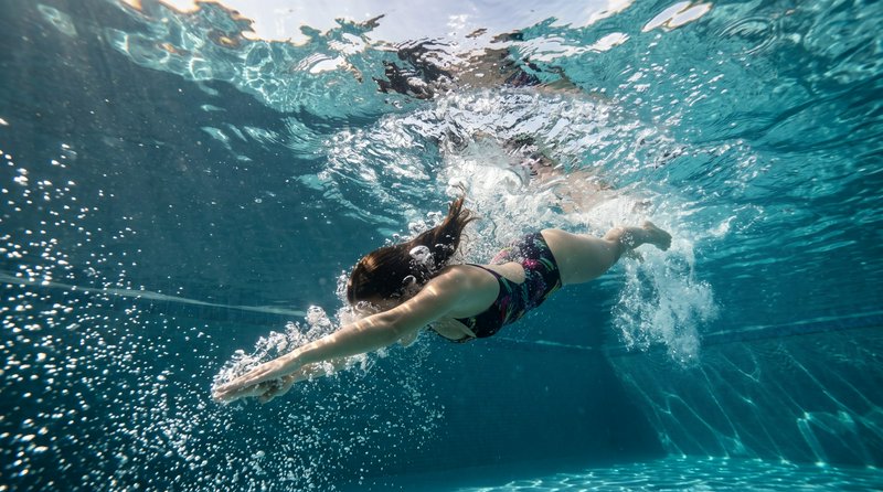 Woman swims forward underwater through clear turquoise pool water surrounded by rising air bubbles