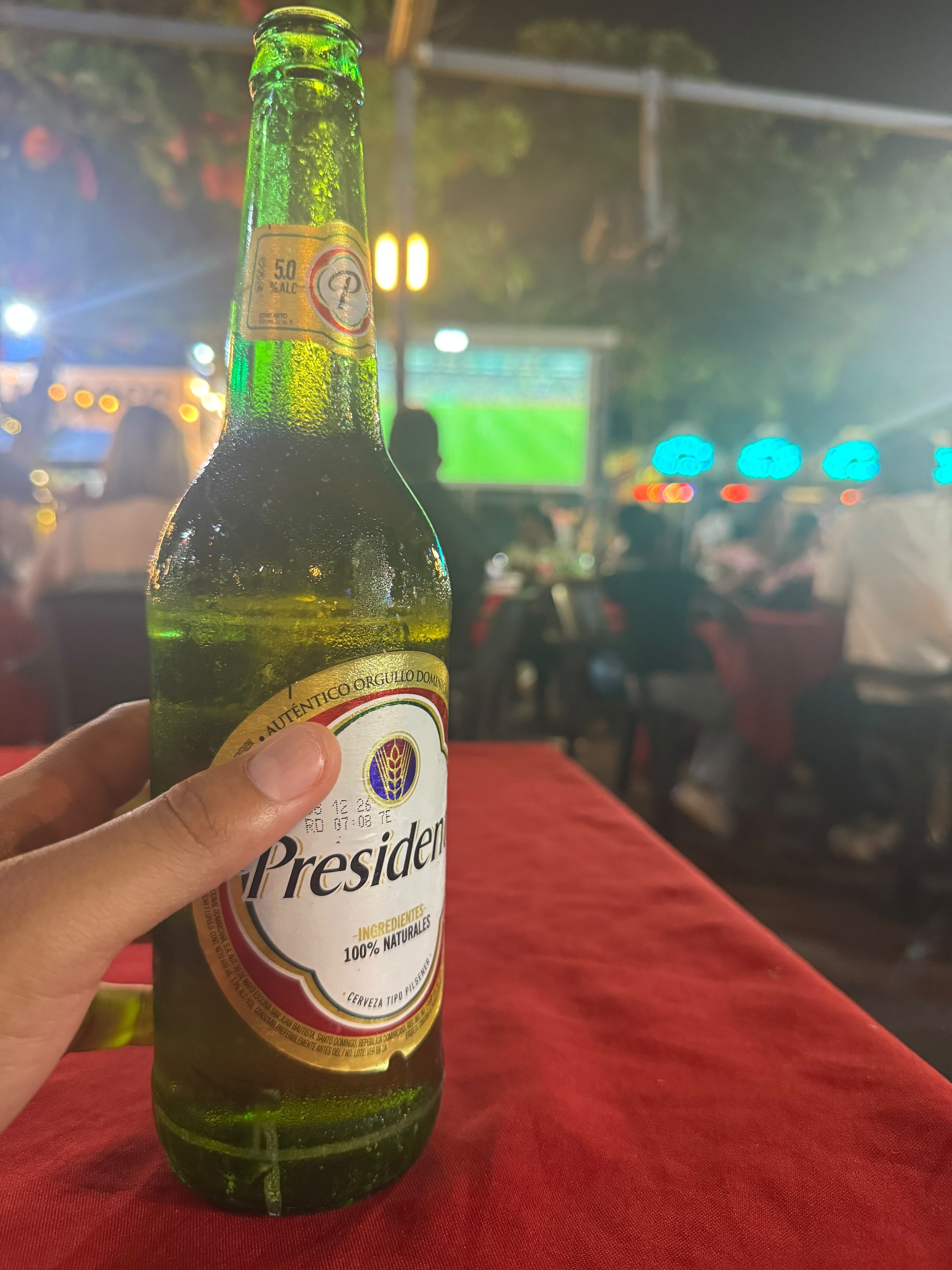 Hand holding a frosty open Presidente beer bottle on a red tablecloth at a lively outdoor night patio bar.