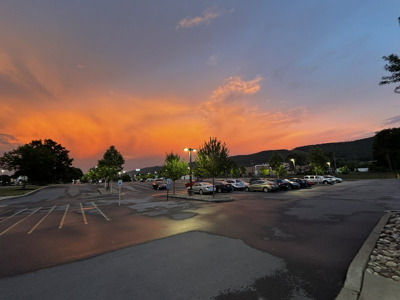 Fiery orange and blue gradient sunset sky over a damp paved parking lot lined with trees and park...