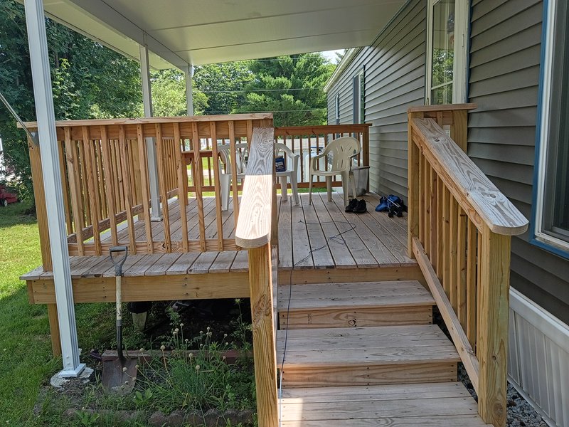 Newly built unfinished wooden residential back porch with stairs, patio chairs, and a tree-lined ...