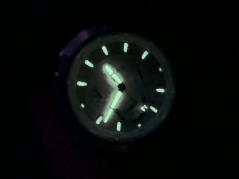 Close up of a luminescent analog watch face glowing pale green against pitch black total darkness.