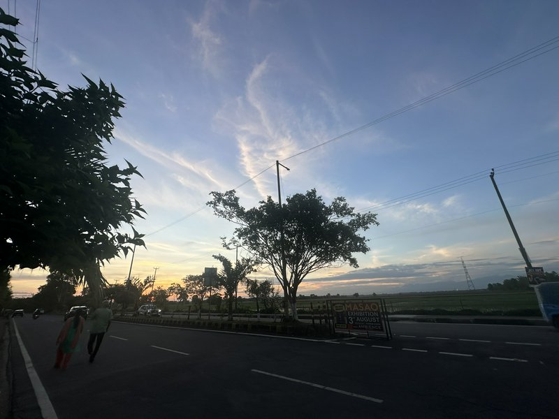 Paved rural roadside at dusk, silhouetted trees, walking pedestrians, soft pastel sunset sky stre...