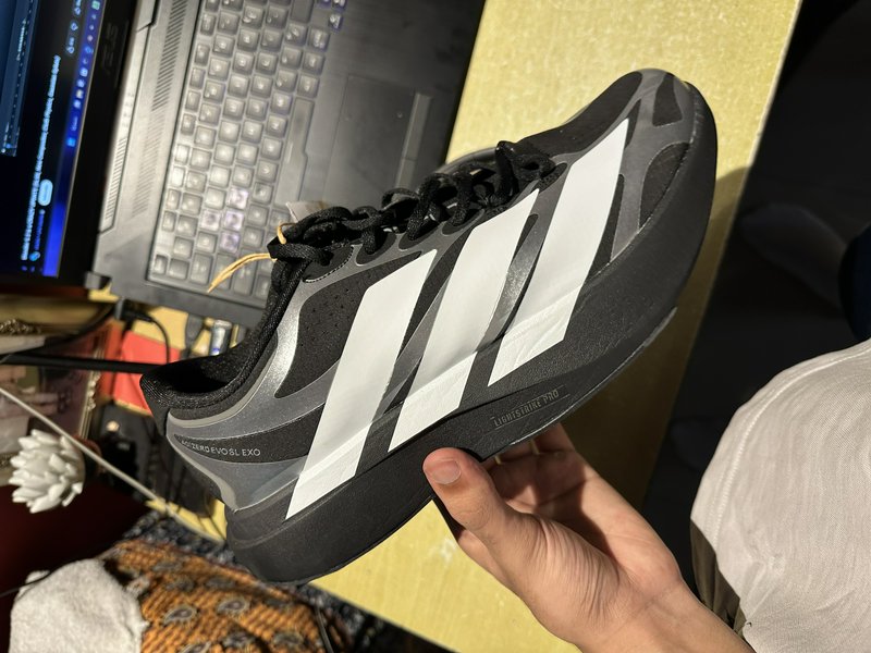 Hand holding a black, white, silver Adidas Adizero Evo SL Exo running shoe above a home desk setup