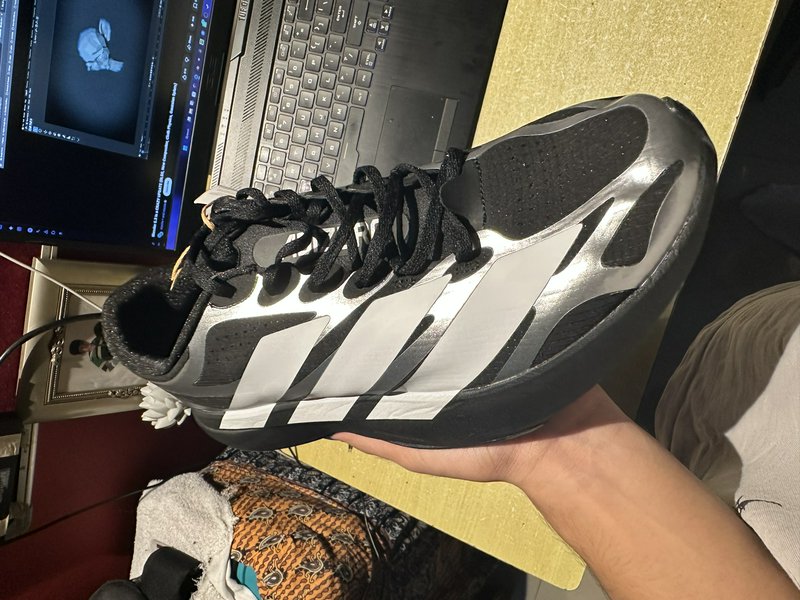 Hand holding a metallic silver and black Adidas running sneaker above a home desk with an open la...