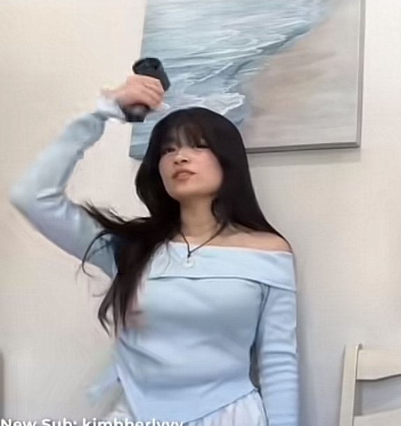 Young woman with long dark hair uses a handheld hair dryer, wearing a pale blue off-shoulder top ...