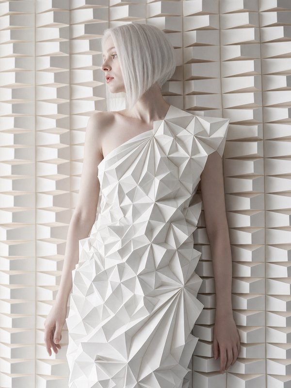 Platinum blonde model in a faceted origami-style white gown stands against a 3D geometric texture...