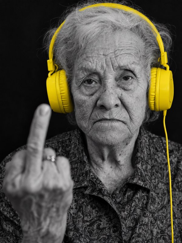 Elderly woman wearing bright yellow headphones gives the middle finger in a selective color studio portrait