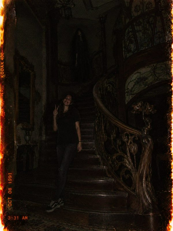 Woman posing on an ornate staircase while a ghostly figure looms on the upper landing above her.