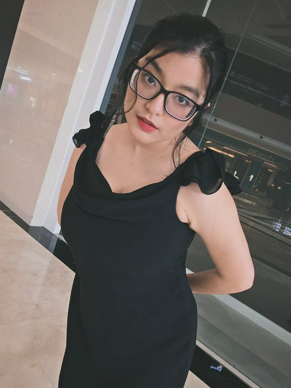 Young woman with black eyeglasses poses in a ruffled shoulder black dress inside a modern shoppin...
