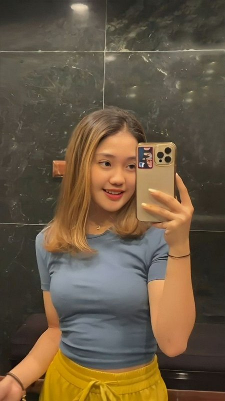 Smiling young woman with highlighted shoulder-length hair takes a mirror selfie wearing a blue to...