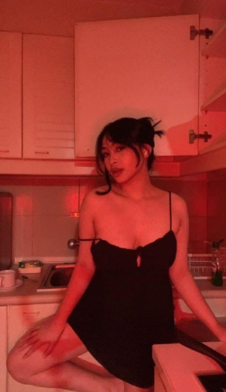Young woman in a black slip dress sits on a kitchen counter bathed in moody red lighting, looking...