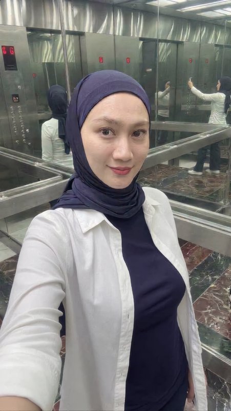 Smiling hijabi woman takes a selfie inside a mirrored stainless steel elevator, wearing a navy he...
