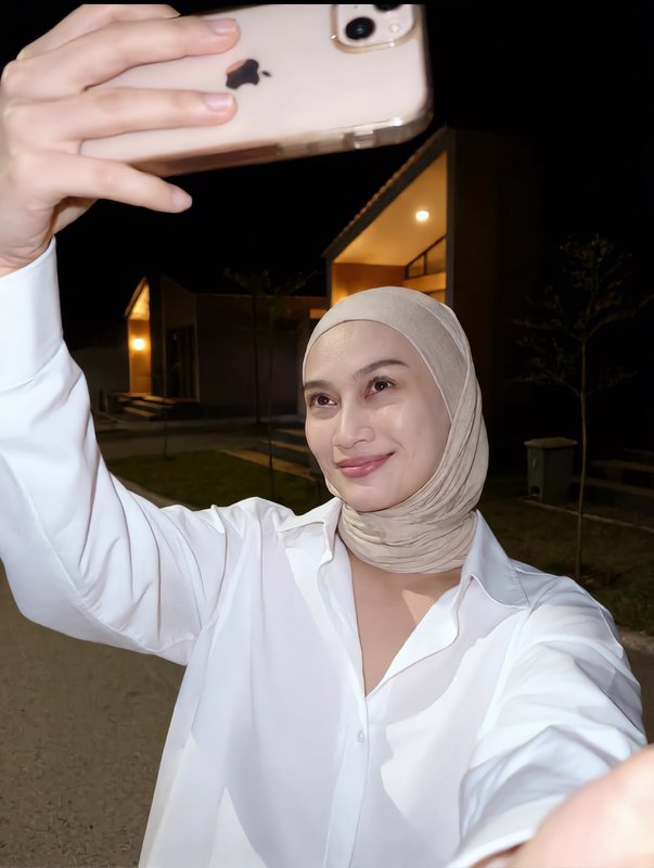 Smiling woman in a beige hijab takes an outdoor selfie with a gold iPhone on a quiet residential ...